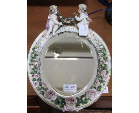 A Continental ceramic mirror frame, cherub with swag mount, floral encrusted, fitted a bevel plate mirror, mirror size 23cm x
