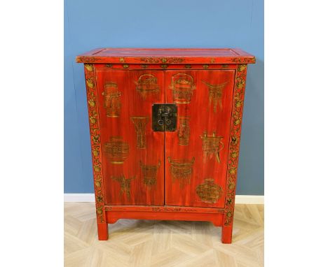 A 20th century red lacquered Chinese two door cabinet of typical form with an interior shelf, decorated with precious objects
