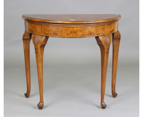 An early 20th century Queen Anne style burr walnut demi-lune fold-over card table with radially veneered top and carved scall