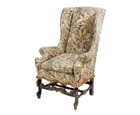 A Queen Anne style wing armchair, upholstered with floral tapestry, on ebonised beechwood scroll front legs, united by a fore