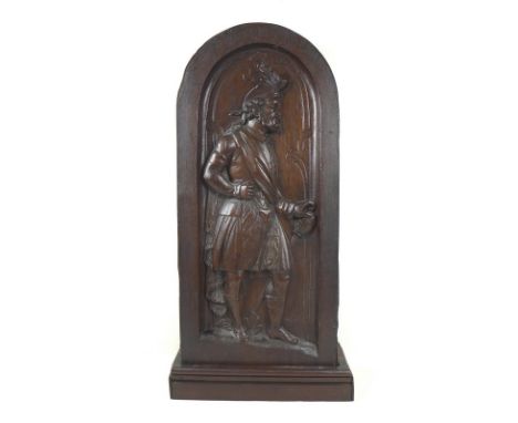 An oak carved panel, 18th / 19th century, depicting a Scottish chieftain with feathered hat, basket hilted sword, kilt and sa