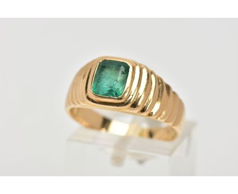 A YELLOW METAL EMERALD SIGNET RING, centring on an emerald cut emerald, bezel set, textured design either side leading onto a