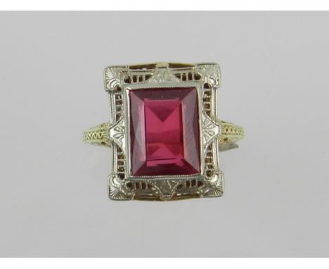 A yellow and white metal Art Deco style dress ring, set with an emerald cut ruby, within an openwork filigree mount.