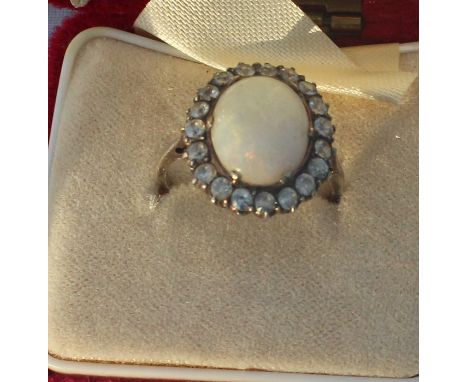 A white metal and opal ring, set oval cabochon precious white opal bordered by round cut clear stones, (white metal assessed 