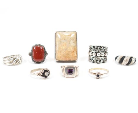 An assortment of silver rings. To include a polished hard stone ring, red stone ring, faceted purple stone ring, two floral w