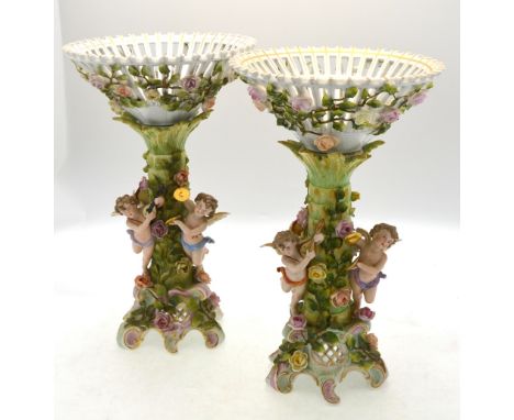 A pair of Sitzendorf porcelain comports, the tall pedestals entwined with climbing roses and supporting three cherubs playing