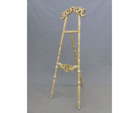 A CREAM PAINTED FLOOR STANDING EASEL, THE FOLIATE BRACKET CRESTING ABOVE TURNED SUPPORTS, THE FLOWER HEAD TOPPED PEGS ADJUSTI