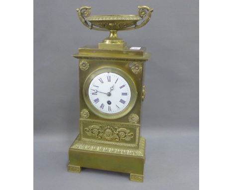 Brass cased mantle clock with a twin handled urn finial over a circular enamel dial with Roman numerals on a rectangular plin