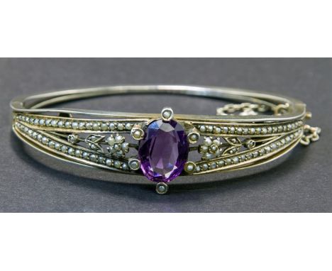 A late 19th century continental amethyst and seed pearl bangle, the central oval amethyst within foliate and plain seed pearl