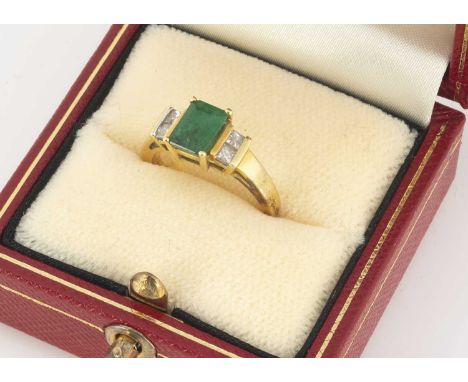 An emerald and diamond dress ring, the rectangular cut stone in a four claw setting, flanked by pairs of princess cut diamond