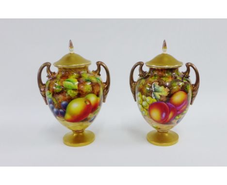 A pair of Royal Worcester floral study lidded urn vases, each with gilded handles and painted with fallen fruit to a mossy gr