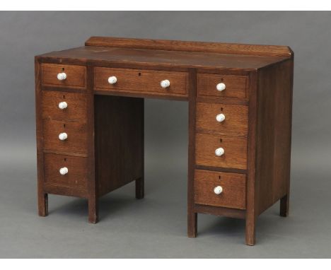 A late Arts and Crafts oak kneehole desk, having a chamfered integral ledgeback, over an arrangement of nine drawers, with tu