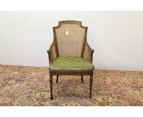 Edwardian mahogany framed bergère library chair with arched caned back and sides and green button leather squab cushion, on s