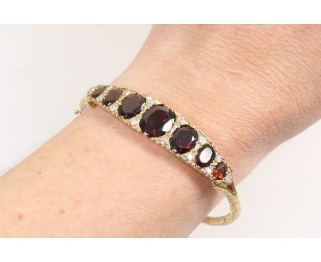 Garnet and diamond hinged bangle in 9ct gold, set with seven graduating oval cut garnets, the largest 10mm x 8mm, dispersed w
