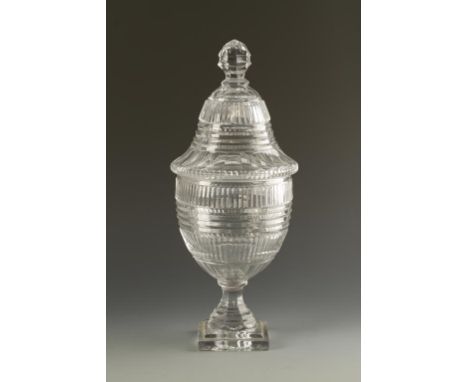 A GEORGE III CUT GLASS URN AND COVER on a waisted and stepped stem and square "lemon squeezer" foot, probably Irish, 17" high