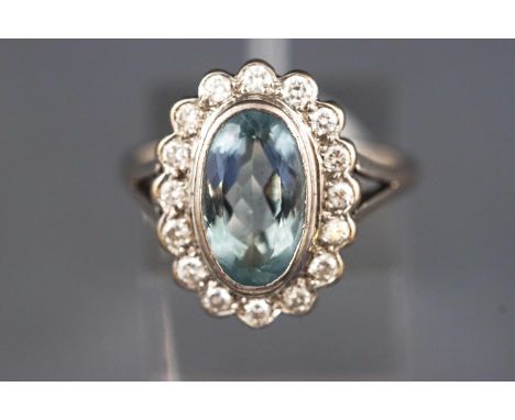 A white metal cluster ring. Set with an oval faceted cut aquamarine and surrounded by round brilliant cut diamonds.  Hallmark