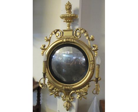 Regency period gilt gesso convex wall mirror with two sconces and flaming urn pediment, scrolled foliate mounts around a circ