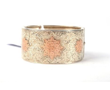 A Victorian cuff bangle, with three applied rose coloured floral motifs, to a foliate chased front, measures 5cm by 6cm inner