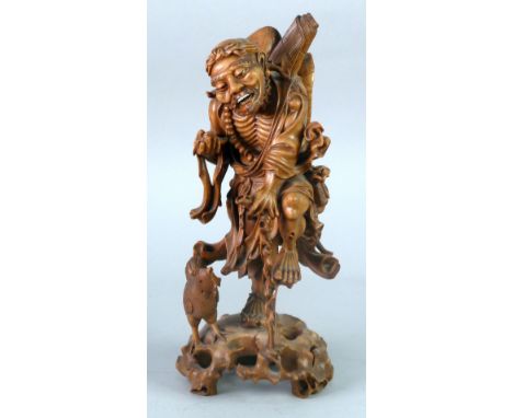 CHINESE CARVED ONE PIECE BLONDE WOOD FIGURE OF A MAN, modelled with one leg raised on a walking stick, carrying a rolled mat 