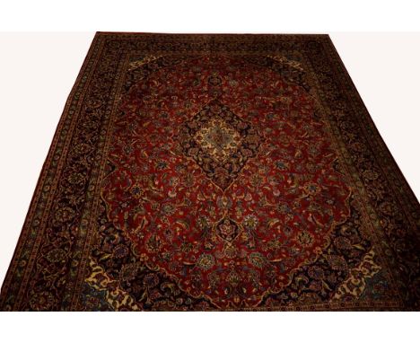 A BURGUNDY GROUND PERSIAN KASHAN CARPET, in all-over floral pattern and centre dark blue medallion, inside a wide blue border