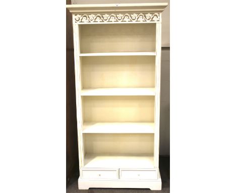 A contemporary wooden bookcase. Painted white with carved details, three shelves over a pair of small frieze drawers, on brac