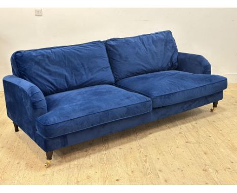 A modern three seater sofa upholstered in deep blue velvet, the feet with brass caps and castors (h.80cm x l. 212cm x w.100cm