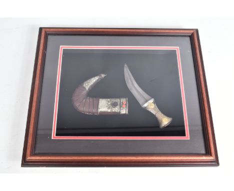 A FRAMED MIDDLE EASTERN STYLE JAMBIYA KNIFE, this has a decorative handle and a curved scabbard, with white metal mounts, the