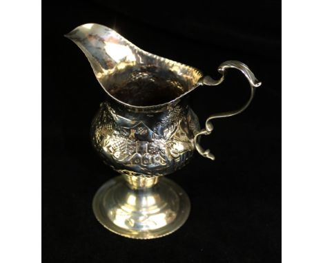 A George II silver cream jugOf bellied helmet form, with an embossed beaded rim and a pastoral scene, to a leaf capped scroll