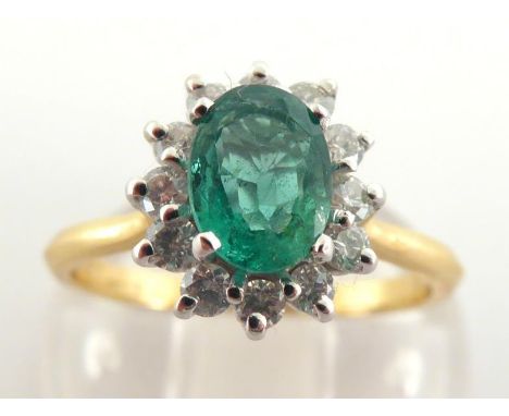 An emerald and diamond cluster ring, the central oval cut emerald 7 x 5mm, in a surround of small brilliants, the shank fully