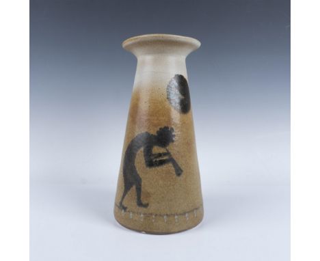 A conical-shaped vase in a cream-khaki finish with salt speckles. Depicts Kokopelli, the flute player, playing under a full m