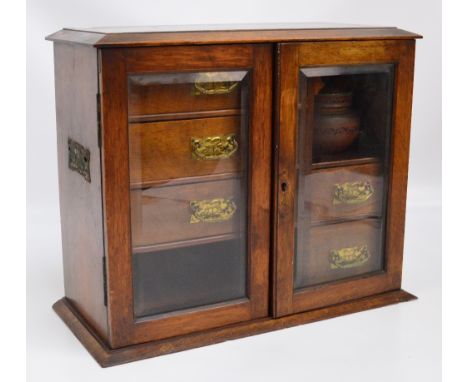 An Edwardian oak smoker's cabinet with a pair of glazed doors enclosing a formation of three drawers above a compartment and 