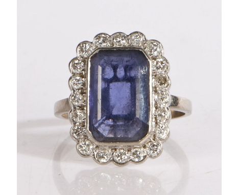 A white metal sapphire and diamond ring, the emerald cut sapphire is bezel set, surrounded by a round brilliant cut diamonds 