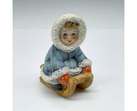 Porcelain figure depicting a child in a blue coat riding a sled. Hand painted in blue, red, and brown, with glass and matte f