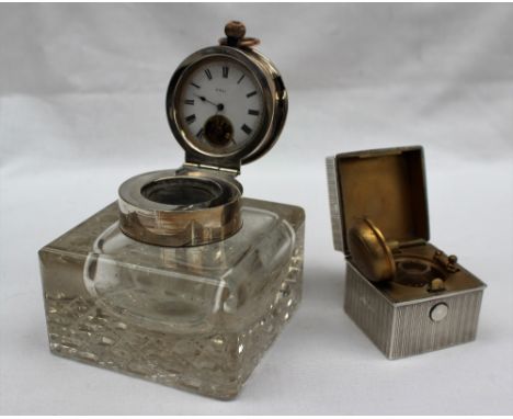 An early 20th century silver topped and glass inkwell, the cover inset with an 8 day pocket watch the enamel dial with Roman 