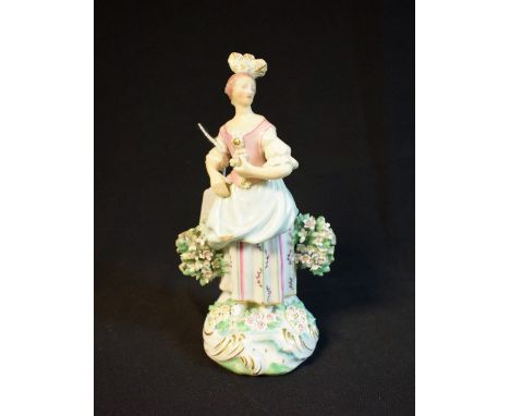 A Derby Patch Mark figure, of a Flower Girl, she stands holding a posy, wearing a feathered headdress, pink bodice, blue skir