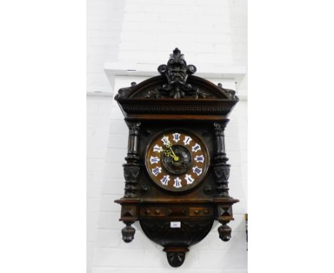 A black forest style wall clock with a carved pediment top with a green man motif to centre over a wooden dial with ceramic n