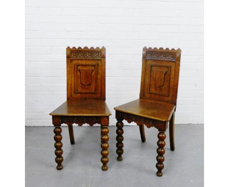 A pair of oak hall chairs in the Gothic style, the carved backs with shield shaped motifs to centre, over solid seats on bobb