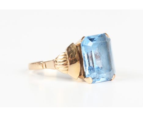 A gold ring, claw set with the cut cornered rectangular step cut synthetic blue spinel between raised shoulders, detailed '75