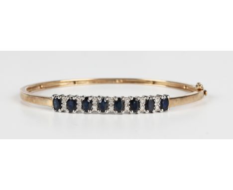 A 9ct gold, sapphire and diamond oval hinged bangle, the front mounted with a row of oval cut sapphires alternating with pair