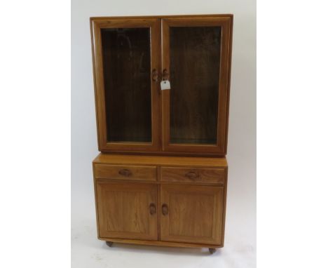 An Ercol light elm cabinet the top section having twin glazed doors enclosing glass shelves, the base with two short drawers 