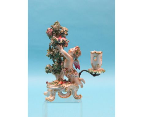 A bow porcelain candlestick figure, cherub youth attempting to catch bird amidst bocage, a torch at his knees, on scroll base