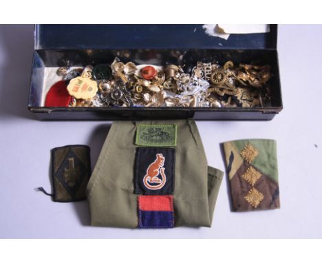 A SMALL BLACK TIN CONTAINING A NUMBER OF PLASTIC CAP BADGES, DEVICES, SHOULDER PIPS, BUTTONS, etc, an Army armband from 1st G