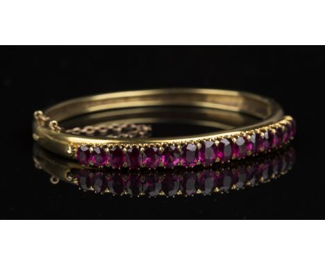 A gold and ruby oval hinged bangle, mounted with a row of fifteen oval cut rubies graduating in size to the centre stone, on 