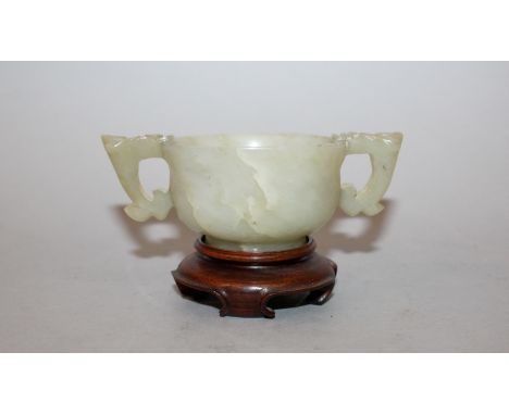 AN EARLY 20TH CENTURY CHINESE JADE TWO-HANDLED CUP, together with a fitted wood stand, the cup handles each in the form of a 