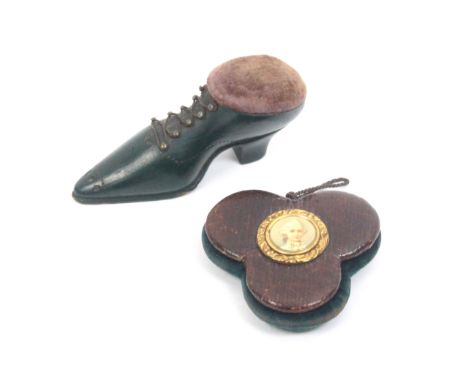 Two pin cushions comprising a carved wooden and green painted example as a lady's shoe, wire lace, 8cm and a leather trefoil 
