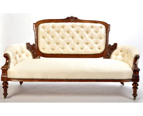 A late Victorian carved walnut settee, the button back, arms and seat covered in cream coloured material surrounded by leaf s