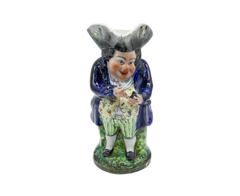 An antique hand painted figural jug of a man in a blue coat, green breeches, yellow floral vest, and a gray tricorn hat. He i