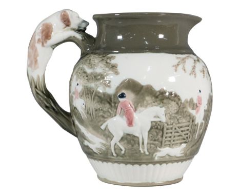 A porcelain pitcher featuring a dog handle and bas-relief hunting scene, lightly colorized in brown, greens, pink, and black 