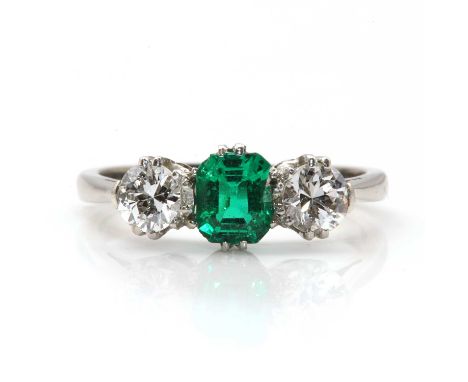 A three stone emerald and diamond ring,with an emerald cut emerald, double claw set to the centre. A brilliant cut diamond, d