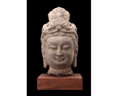 Ca. 581-618 AD. A stone head of Buddha with elaborate coiffured hair curls under a richly ornamented headdress. He is charact